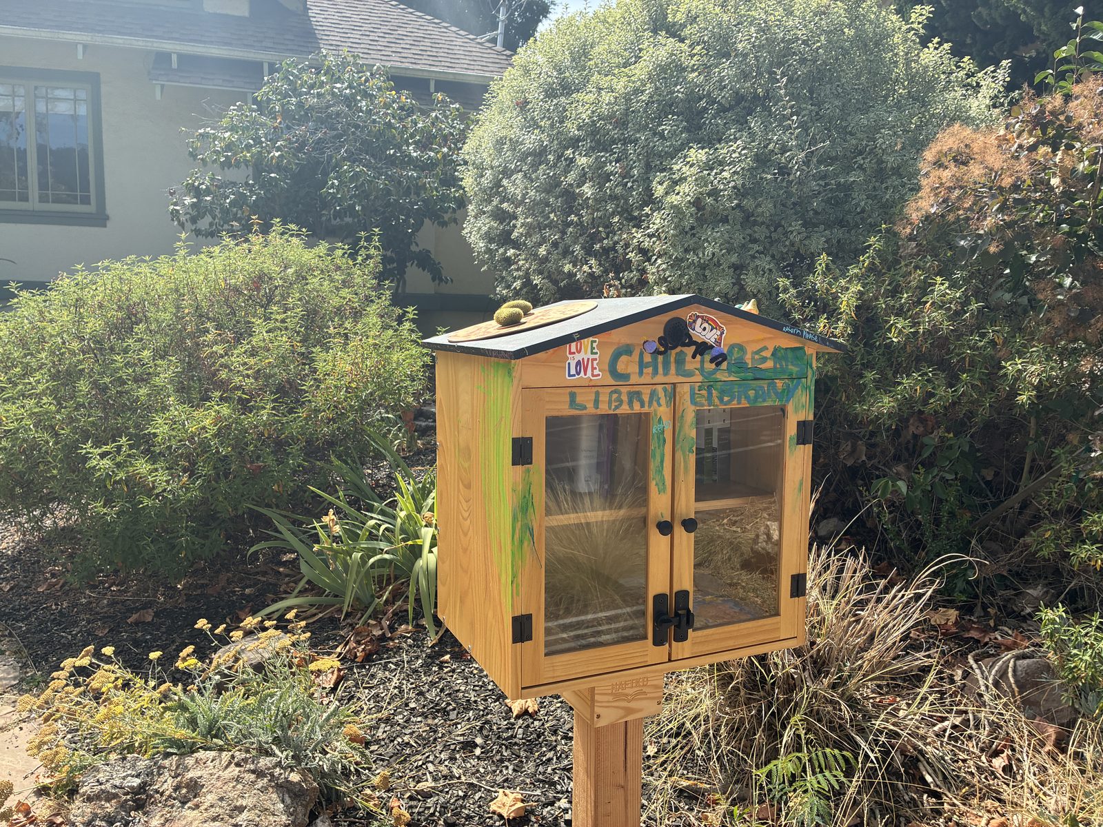 A Little Free Library box on a post, half hidden in the shrubs