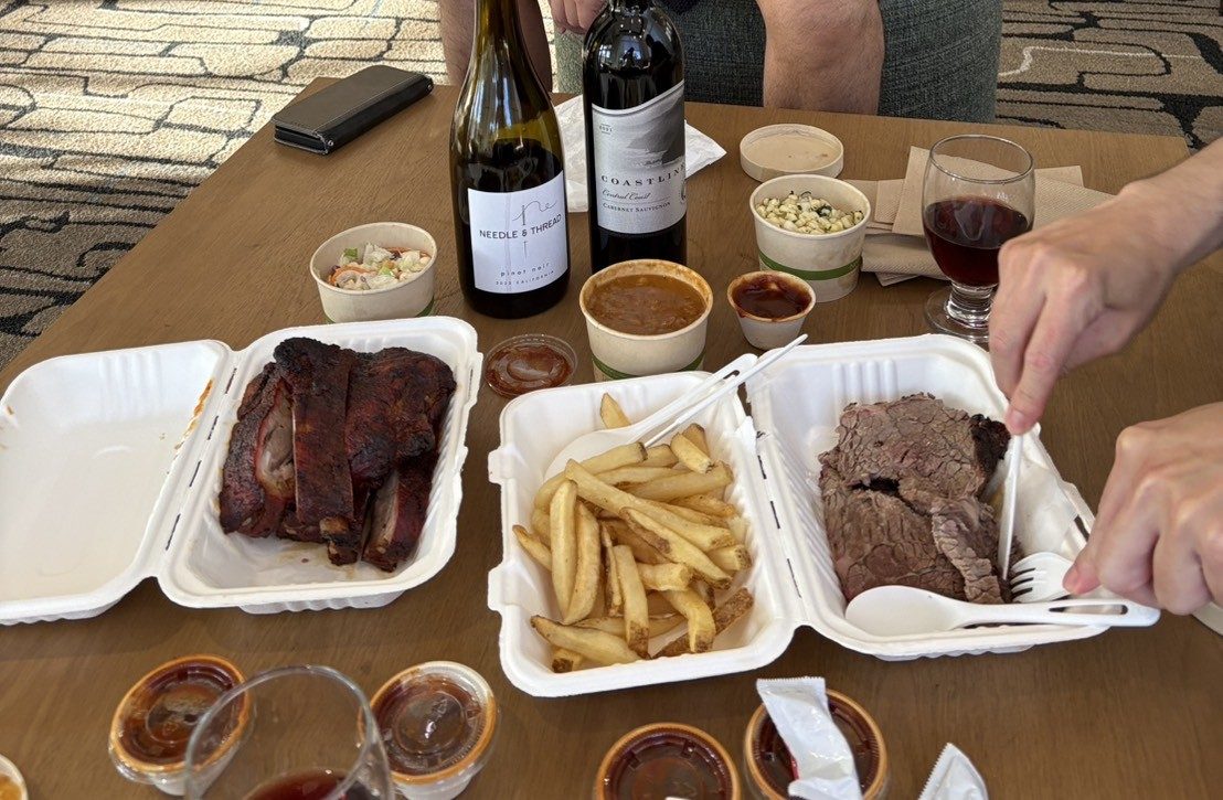 Takeout containers of ribs, fries and brisket spread on a table, a bottle of red wine alongside