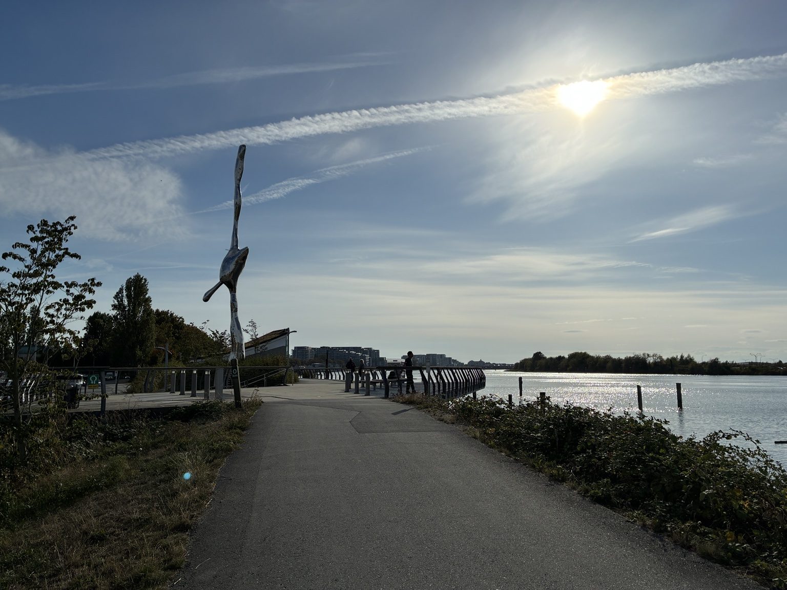 水面に日が差す川沿いの遊歩道。手すりのそばに背の高い彫刻がある